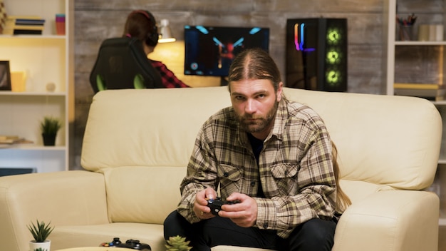 Image of a gamer sitting comfortably on a sofa, intensely focused on a PC game displayed on a large monitor, holding a wireless gaming controller in their hands. The lighting in the room is dim, enhancing the immersion.
