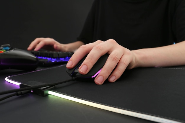 A close-up of a gamer's hands on a gaming keyboard and mouse, highlighting the ergonomic setup and the focus on precision and control. The background is softly blurred, showing a high-end gaming PC with colorful RGB lighting.