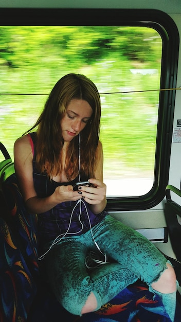 A user holding the Nintendo Switch OLED while sitting on a train, playing a colorful Nintendo game. The focus is on the screen and the user's hands. The background shows the blurred scenery outside the train window.