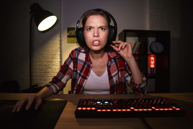 A person adjusting the microphone of their gaming headset while sitting at a computer, with a second monitor displaying a game in the background.